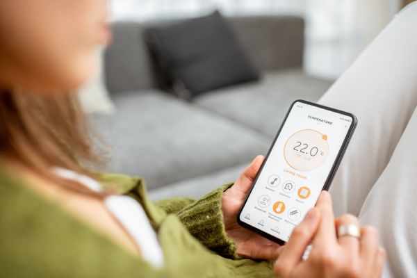Frau hält ein Smartphone in der Hand, auf dem eine App für die Smart-Home-Steuerung angezeigt wird.