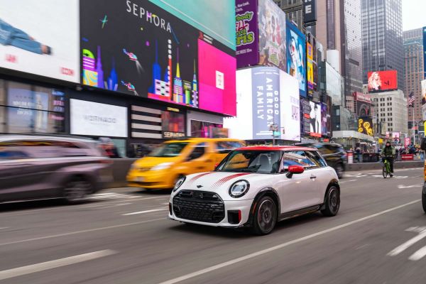 Ein weißer Mini JCW mit rotem Dach fährt durch eine Großstadt mit Wolkenkratzern.