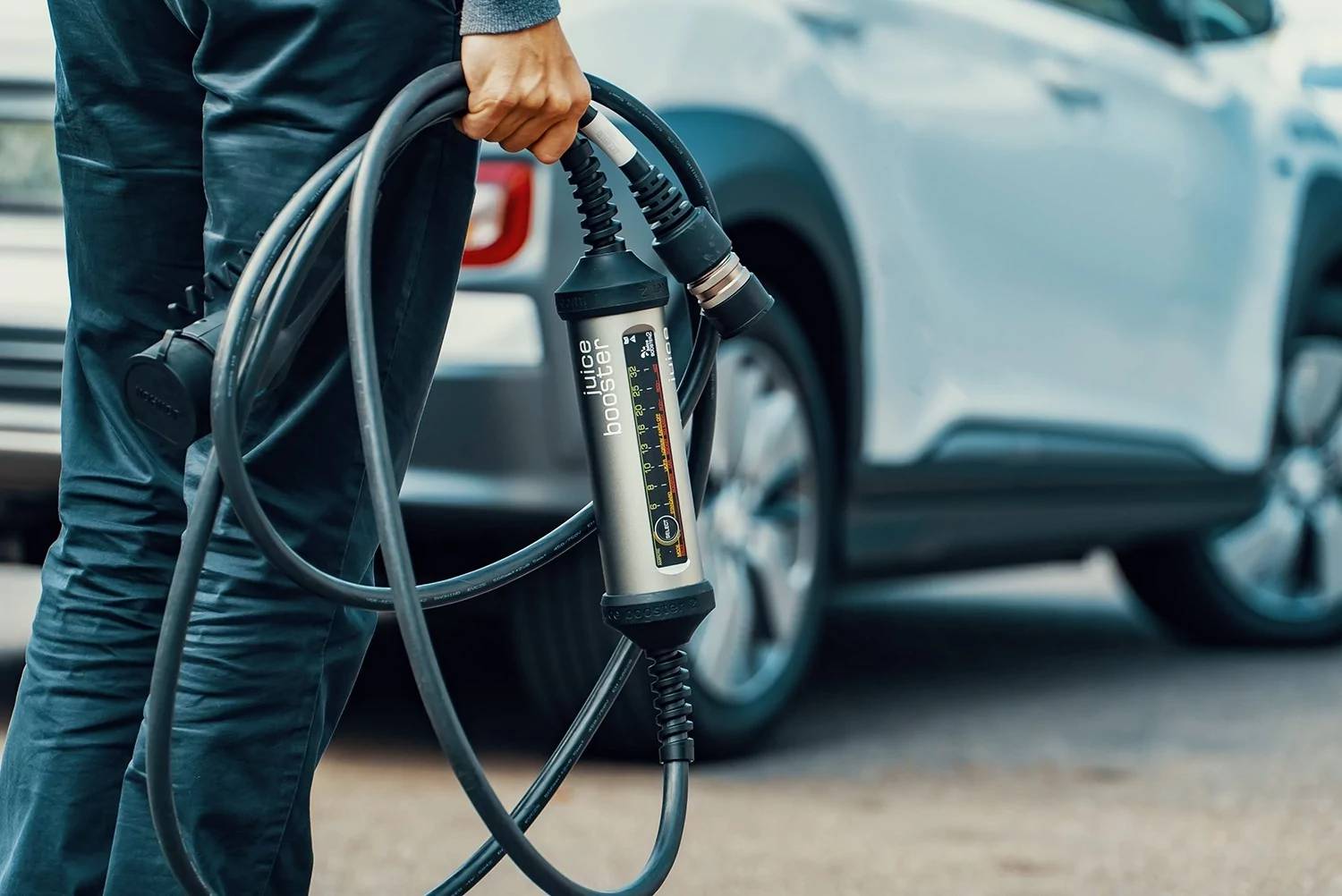 Ein Mann trägt ein Ladekabel für ein E-Auto, Fahrzeug im Hintergrund. 