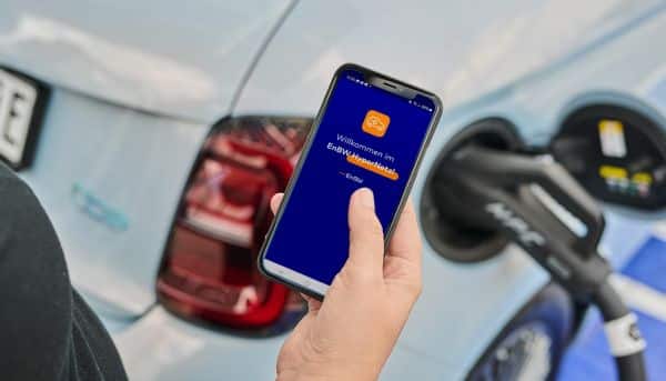 Hand hält ein Smartphone, auf dem die EnBW mobility+ App zu sehen ist, im Hintergrund ein E-Auto, das geladen wird.